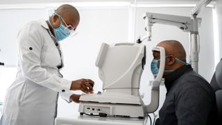 a Black middle-aged man wearing a surgical mask sits in an examination chair in front of an optometry device as a doctor examines his eyes
