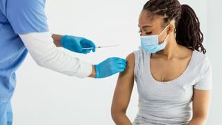 Person wearing a blue face mask sits and watches a nurse who is preparing their arm for vaccination