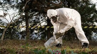 Forensic scientist in a white plastic suit collects a pair of shoes in a plastic bag in a taped off crime scene in the woods